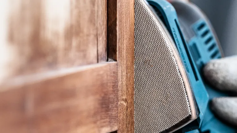 Een persoon met beschermende uitrusting schuurt een houten deur met een detailsschuurmachine.