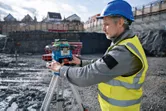 Een persoon met veiligheidsuitrusting stelt een laserwaterpas af op een statief op een bouwterrein.
