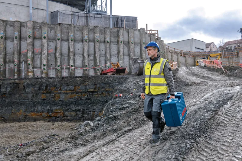 Een persoon die veiligheidsuitrusting draagt, draagt een blauwe gereedschapskoffer op een modderige bouwplaats.