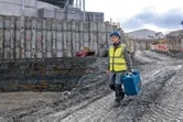 Een persoon die veiligheidsuitrusting draagt, draagt een blauwe gereedschapskoffer op een modderige bouwplaats.