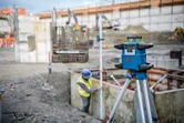 Een laserwaterpasapparaat is opgesteld op een bouwterrein; een persoon met veiligheidsuitrusting meet de diepte.