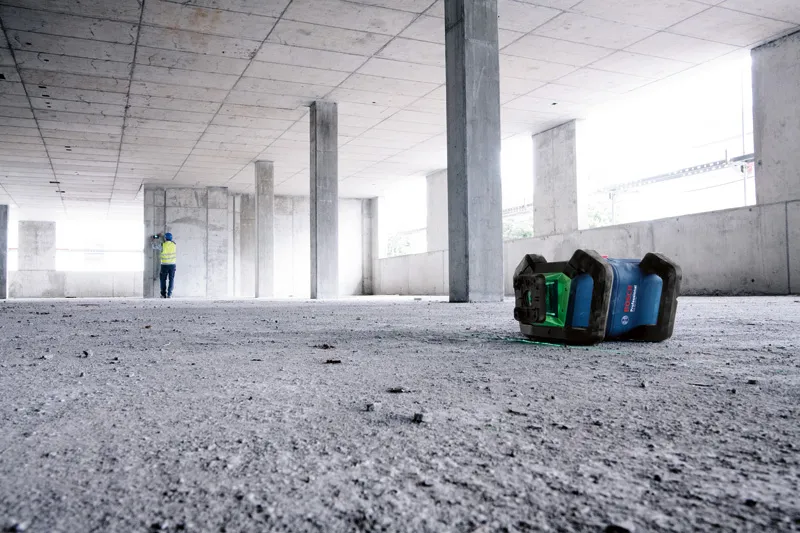 Een laserwaterpas projecteert een groene lijn op een betonnen gebouw terwijl een persoon met veiligheidsuitrusting aan het werk is.