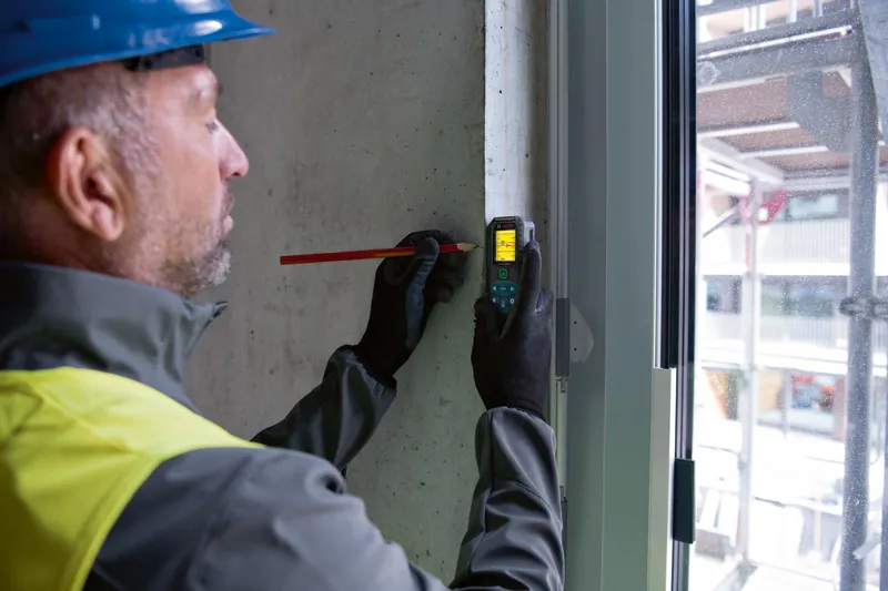 Een persoon die veiligheidsuitrusting draagt, gebruikt een lasermeter om een meting op een betonnen muur te markeren.