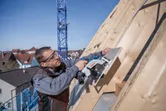 Een persoon met veiligheidsuitrusting zaagt houten planken op een schuin dak met een cirkelzaag.
