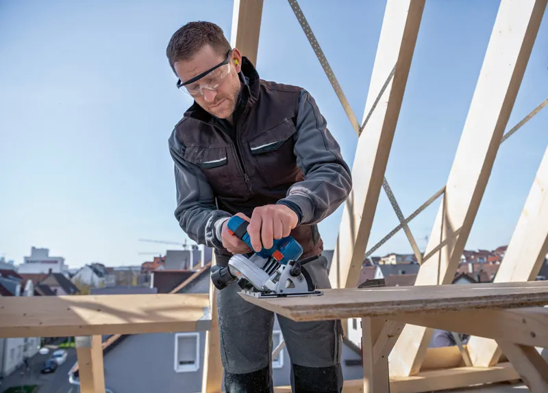 Een persoon met veiligheidsuitrusting zaagt hout met een snoerloze cirkelzaag op een bouwplaats.