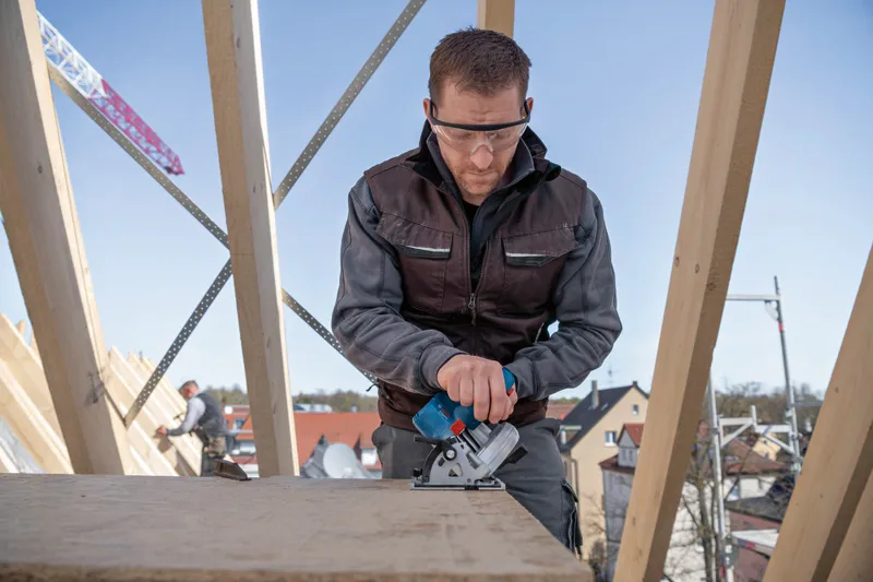 Een persoon die veiligheidsuitrusting draagt, gebruikt een snoerloze cirkelzaag op een houten plank.