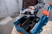 Iemand plaatst een draadloze cirkelzaag in een blauwe gereedschapskoffer op een werkbank.