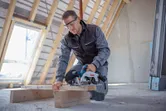 Een persoon met veiligheidsuitrusting zaagt een houten plank door met een draadloze cirkelzaag.