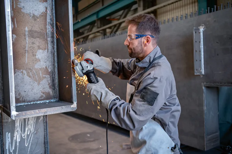 Een persoon met veiligheidsuitrusting slijpt de rand van een grote metalen balk.