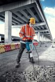 Een persoon met veiligheidsuitrusting breekt buitenshuis beton met een grote elektrische breekhamer.