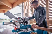Iemand die veiligheidsuitrusting draagt, gebruikt een verstekzaag om een ​​houten balk door te zagen.