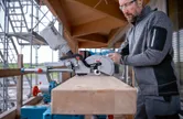 Een persoon die veiligheidsuitrusting draagt, stelt een verstekzaag af om een ​​houten balk door te zagen.