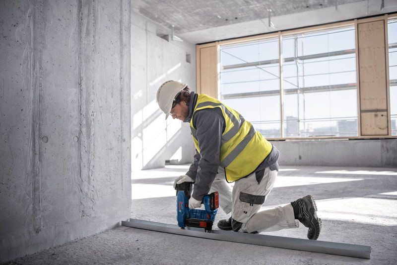 Een persoon die veiligheidsmateriaal draagt, bevestigt met een draadloze betonspijkermachine een metalen spoor aan de vloer.