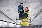Een persoon met veiligheidsuitrusting boort met een elektrisch gereedschap in een betonnen plafond.
