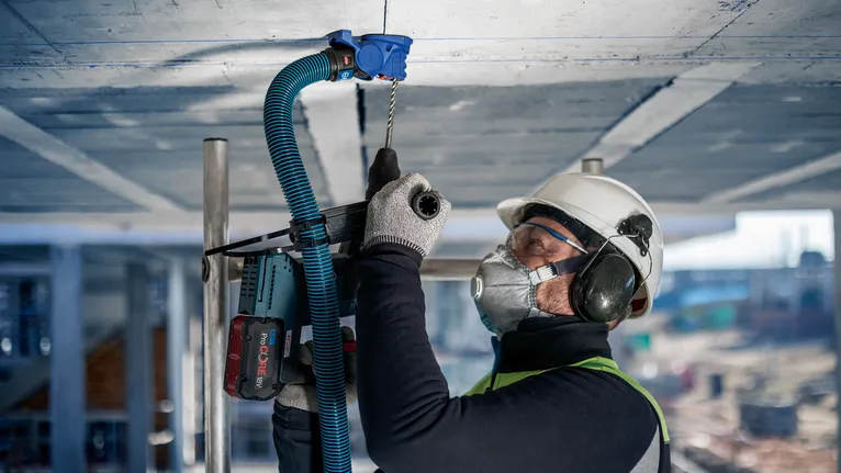 Een persoon met veiligheidsuitrusting boort in een betonnen plafond op een bouwplaats.