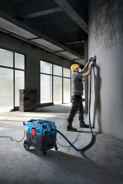 Een persoon met veiligheidsuitrusting slijpt een betonnen muur met behulp van een alleszuiger.