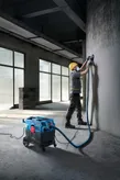 Een persoon met veiligheidsuitrusting slijpt een betonnen muur met behulp van een alleszuiger.