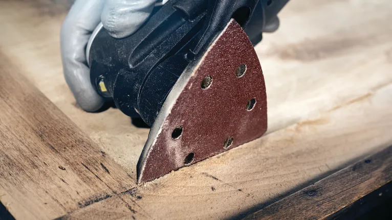 Een persoon die veiligheidsuitrusting draagt, schuurt een houten oppervlak met een detailschuurmachine.