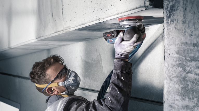 Een persoon met veiligheidsuitrusting gebruikt een elektrische schuurmachine op een betonnen plafond.