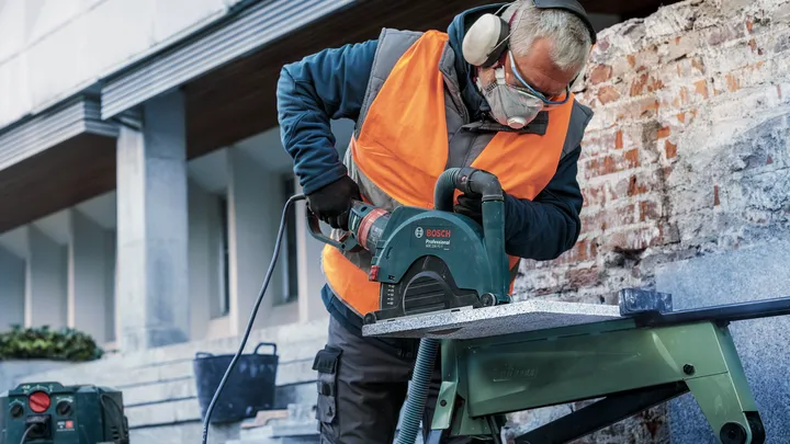 Een persoon met veiligheidsuitrusting zaagt stenen met een cirkelzaag op een werkbank.