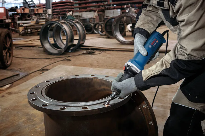 Een persoon met veiligheidsuitrusting slijpt de rand van een grote metalen cilinder met een rechte slijper.