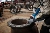 Een persoon met veiligheidsuitrusting slijpt de rand van een grote metalen cilinder met een rechte slijper.