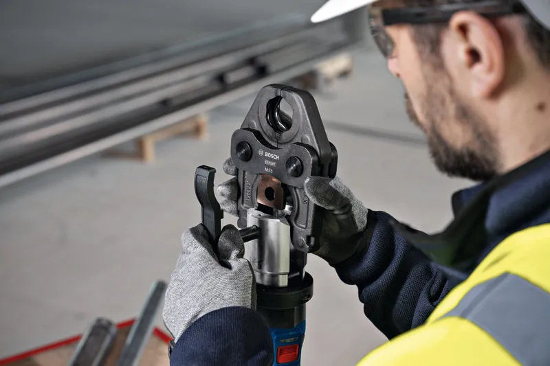 Een persoon die veiligheidsuitrusting draagt, bedient een snoerloos persgereedschap op een metalen fitting.