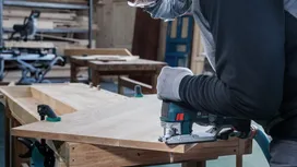Een persoon met veiligheidsuitrusting zaagt hout met een decoupeerzaag in een werkplaats.