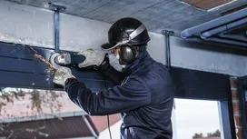 Een persoon met veiligheidsuitrusting slijpt met een elektrisch gereedschap een metalen balk boven zijn hoofd.