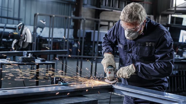 Een persoon met veiligheidsuitrusting slijpt in een werkplaats een metalen balk, waarbij vonken ontstaan.