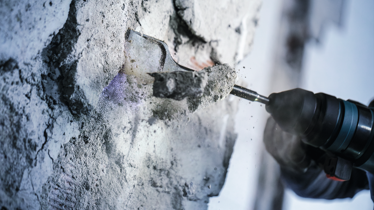 Een persoon met veiligheidsuitrusting beitelt beton uit een muur met een boorhamer.