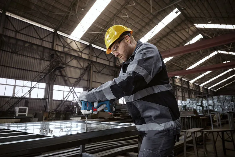 Een persoon die veiligheidsuitrusting draagt, gebruikt een boormachine op een metalen frame in een fabriek.