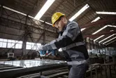 Een persoon die veiligheidsuitrusting draagt, gebruikt een boormachine op een metalen frame in een fabriek.