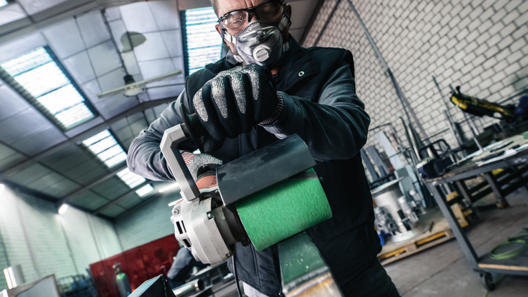 Een persoon die veiligheidsuitrusting draagt, bedient een elektrisch gereedschap met een groene schuurrol in een werkplaats.