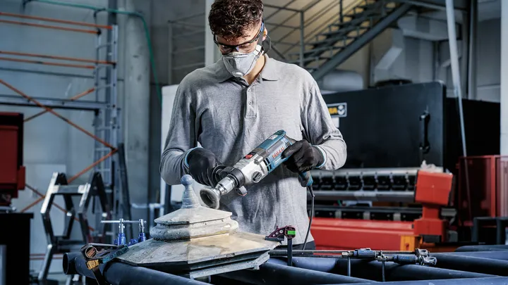 Een persoon die veiligheidsuitrusting draagt, polijst een metalen constructie met een elektrisch gereedschap.