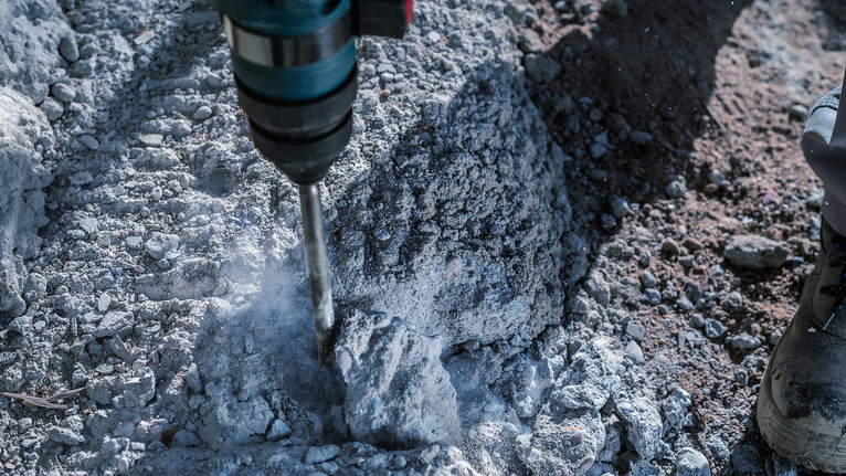 Een boorhamer slaat puin van beton in stukken terwijl een persoon veiligheidsuitrusting draagt.