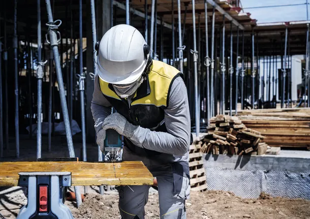 Een persoon met veiligheidsuitrusting zaagt op een bouwplaats een houten plank met een elektrisch gereedschap.