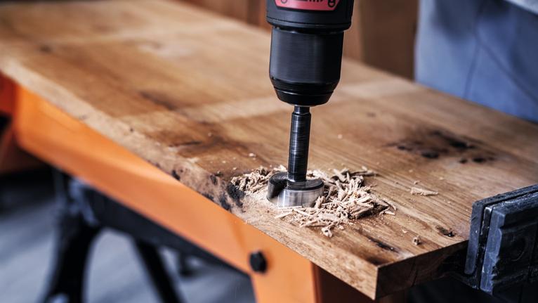 Iemand gebruikt een boormachine met een gatenzaag om door een houten plank te zagen.