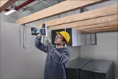 Een persoon die veiligheidsuitrusting draagt, gebruikt een boormachine om metalen steunen boven zijn hoofd te bevestigen.