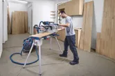 Een persoon die veiligheidsuitrusting draagt, zaagt in een werkplaats een houten plank met een verstekzaag.