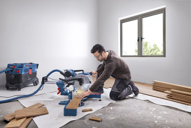 Een persoon met veiligheidsuitrusting zaagt houten planken met een verstekzaag in een lichte kamer.