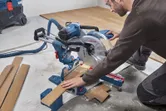 Een persoon met veiligheidsuitrusting zaagt een houten vloer met een verstekzaag.