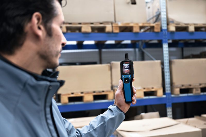 Iemand gebruikt een thermo-hygrometer in een magazijn met planken vol dozen.