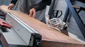 Persoon duwt een groot houten bord door een tafelzaag in een werkplaats.