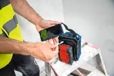 Een persoon die veiligheidsuitrusting draagt, laadt zijn telefoon op met een draagbare powerbank terwijl hij op een ladder staat.
