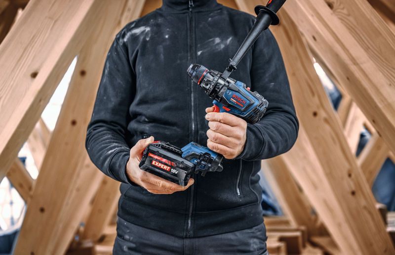 Een persoon houdt een accuboormachine en een accu vast in een houten constructieframe.