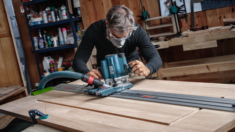 Een persoon met veiligheidsuitrusting leidt een freesmachine langs een spoor om een houten plank in vorm te brengen.