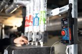 Een persoon inspecteert leidingen terwijl een digitale multimeter in de buurt de temperatuur meet.