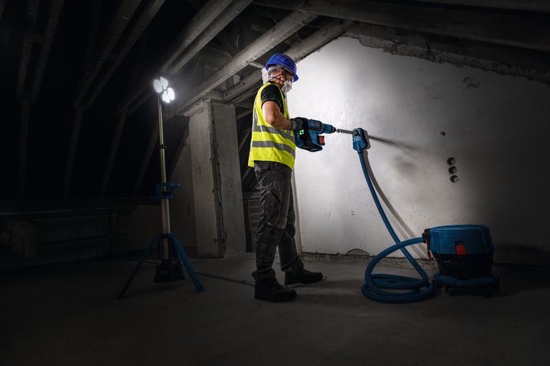 Een persoon met veiligheidsuitrusting boort in een muur onder een draadloze bouwlamp.