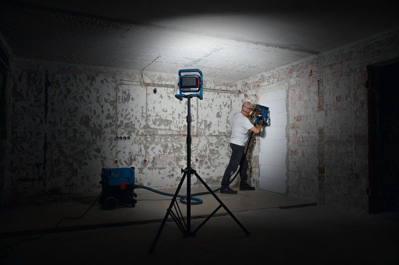 Een persoon die veiligheidsuitrusting draagt, bedient een muurboorgereedschap onder een werklamp.